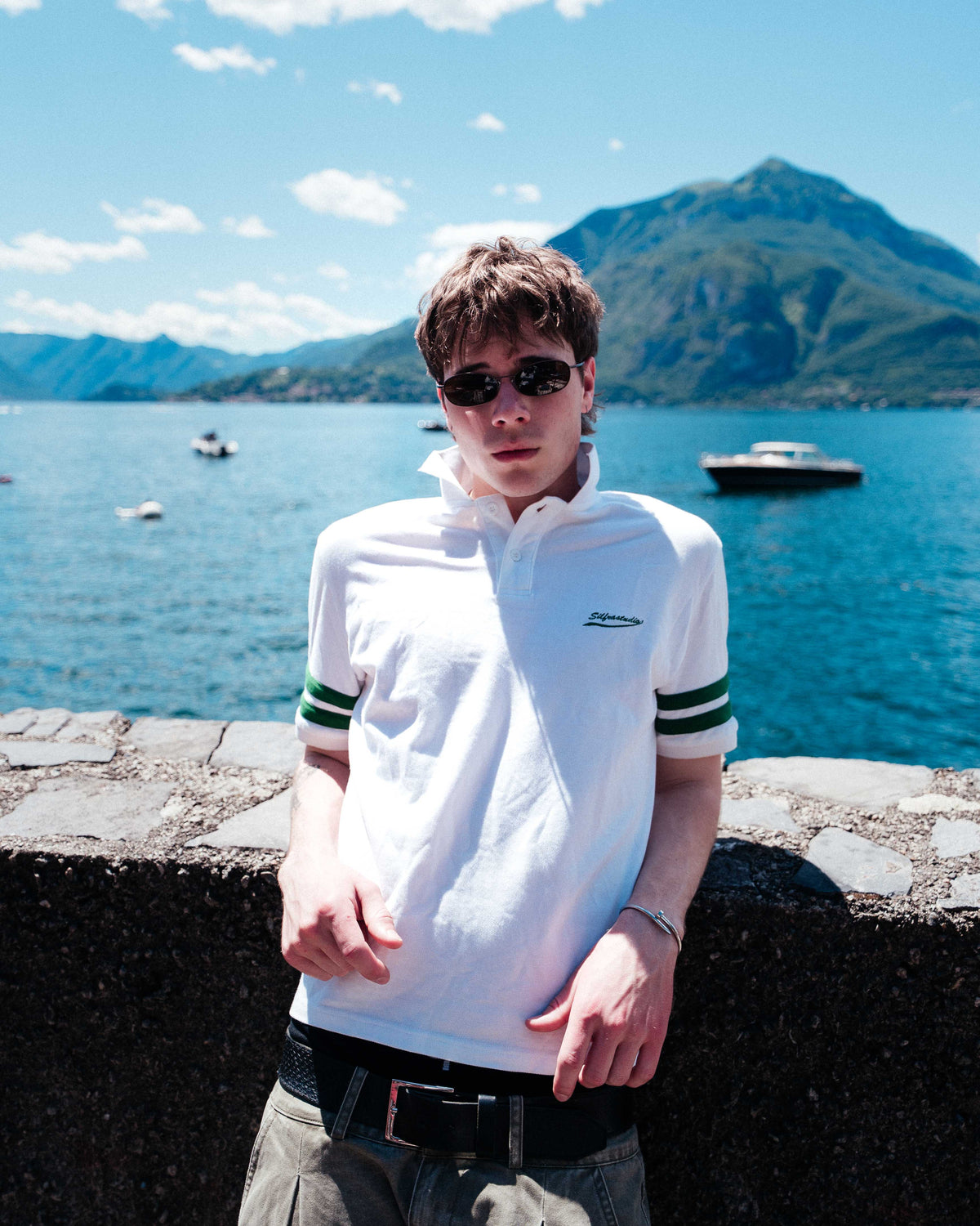 A man wearing a white polo shirt with green stripes on the sleeves, sunglasses, and a black belt stands outdoors in front of a scenic lake and mountain background.
