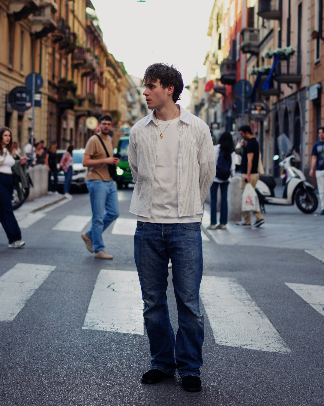 A man standing on a city street crosswalk wearing a light beige button-up shirt over a white t-shirt, blue jeans, and black shoes, with blurred pedestrians and buildings in the background.
