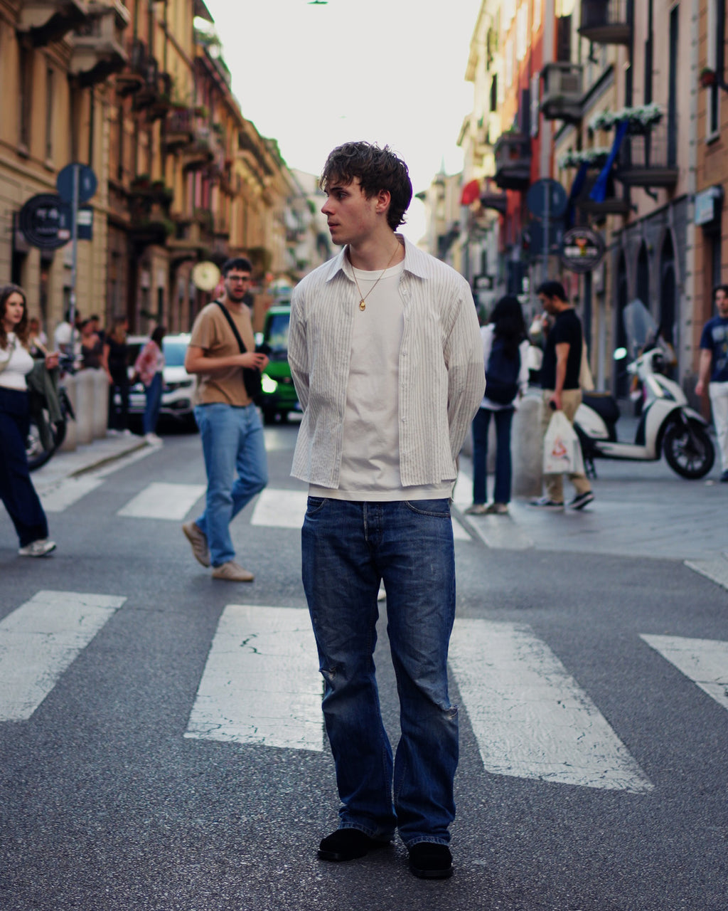 A man standing on a city street crosswalk wearing a light beige button-up shirt over a white t-shirt, blue jeans, and black shoes, with blurred pedestrians and buildings in the background.