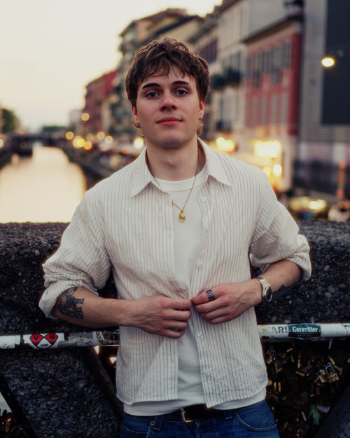 A young man with short brown hair stands on a bridge by a canal at dusk, wearing a cream-colored ribbed button-up shirt over a white t-shirt and blue jeans.